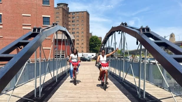 A mother and daughter happily riding bikes over a wooden  bridge at Buffalo's Canalside under a clear blue sky. The bridge has dark arches, and brick buildings are visible in the background.