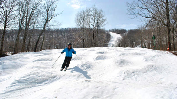 A skier in a blue jacket and black pants skiing along a bumpy snowy slope on Catamount Mountain