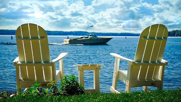 Two white deck chairs and a small wooden table sit in front of Chautauqua Lake on a sunny day with a boat cruising past in front of them