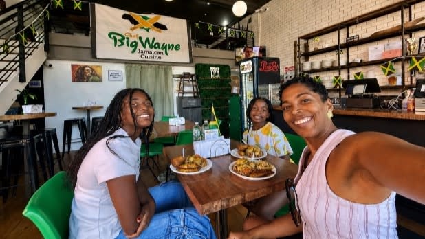 Three people smiling at a table inside "Big Wayne Jamaican Cuisine." They sit in a bright, casual eatery, enjoying plates of Jamaican food.