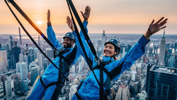 Two people in blue jumpsuits leaning out over the edge of Edge NYC in harnesses with views of the skyline in the background