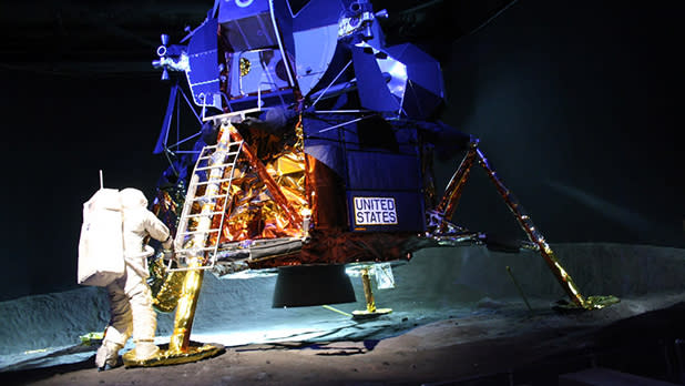 Astronaut in a white suit stands near a replica of the lunar module, labeled "United States," on a moon-like surface, conveying a sense of exploration.