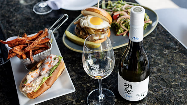 A bottle of sake, a glass, a lobster roll with fries, and a burger with a salad on a table