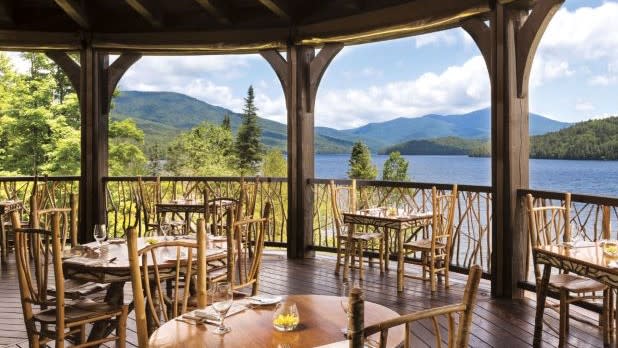 Rustic wood table and chairs on a covered outdoor patio looking out at Mirror Lake at Artisans Restaurant at the Lake Placid Lodge