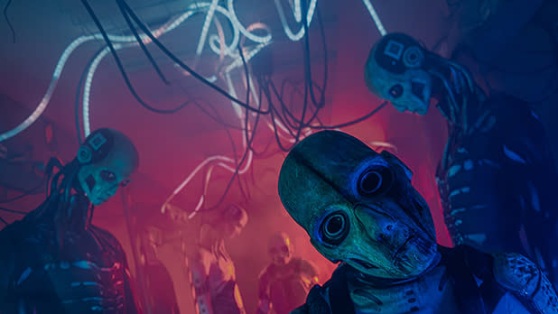 A creepy character with huge dark eyes looms in the foreground as two more figures stand beneath tangled cables in a red-lit, eerie sci-fi scene at Frightworld America's Screampark.