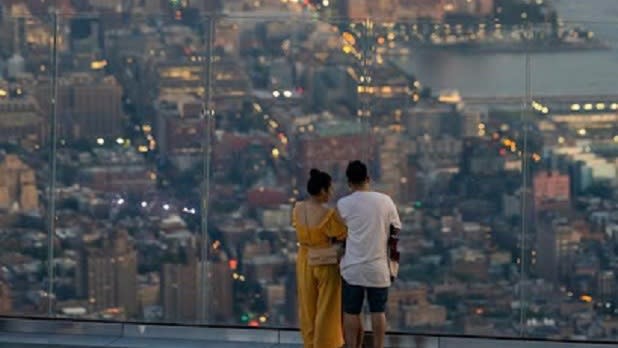 A woman in a yellow jumpsuit and a man in a white t-shirt and black shorts lookout at the NYC skyline at dusk
