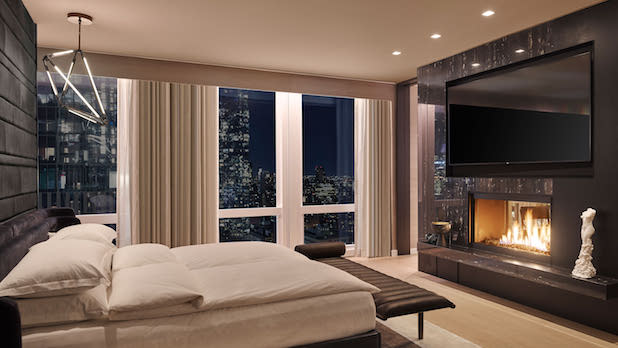 Interior of a luxurious bedroom with a fireplace, large flat screen TV, comfortable bed, and large floor to ceiling windows with a city view in the background