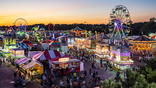 An aerial view of the bustling Erie County fairgrounds at sunset with colorful tents, food stands, carnival rides, bright lights, and crowds of people.