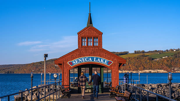 A couple walks towards a charming red boathouse with a steeple on Seneca Lake pier. The sky is clear, and autumn foliage colors the distant hills.