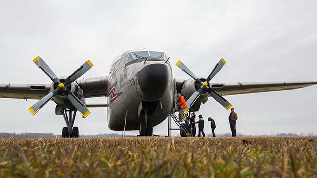 A large, propeller-driven military aircraft is parked on a field. Several individuals are inspecting it, with one man standing on a ladder near its open hatch.