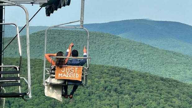 A bride and groom sit on a ski lift that reads "Just Married" on the back while looking out over the Catskill Mountains