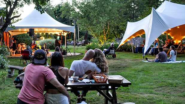 Two couples sit at a picnic table looking out at a stage under a white tent