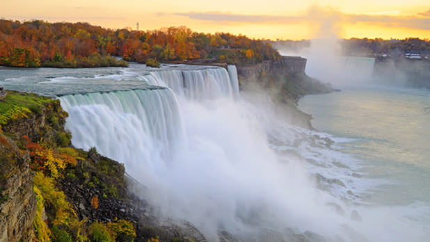 Water rushing down Niagara Falls and mist rising back up with orange, yellow and green trees in the distance