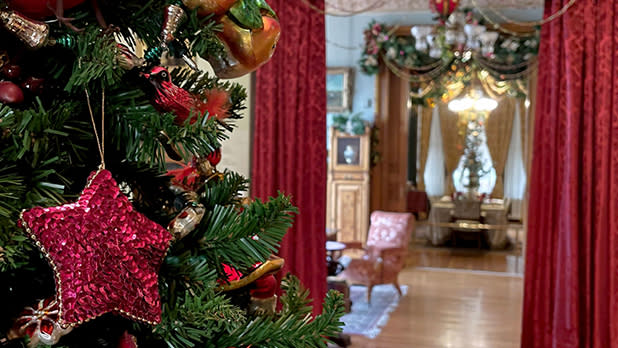Christmas tree close-up shows shiny red star and bird ornaments, set in an opulent, festive room with red curtains, garlands, and elegant decor at the Hudson River Museum's historic Glenview home.