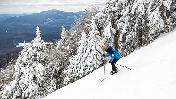 A skier in a blue jacket and black pants skiing down a steep snowy slope past snow-covered trees on Gore Mountain in the Adirondacks