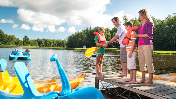 two parents and two kids with in life jackets and paddles stand on a dock on a lake, preparing to go out on boat.
