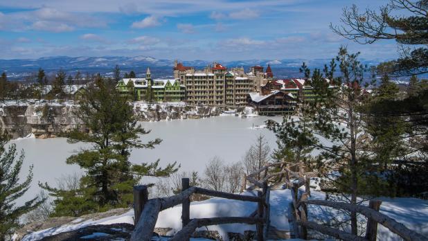 A large hotel complex sits atop a cliff overlooking a frozen lake, surrounded by snow-covered trees. The sky is partly cloudy.
