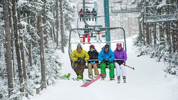 Groups of people are sitting in ski lifts traveling along the snowy tree-lined slopes at Holiday Valley