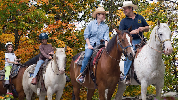 A Rocking Horse Ranch guide on horseback leads an adult and two kids on horses through fall foliage views