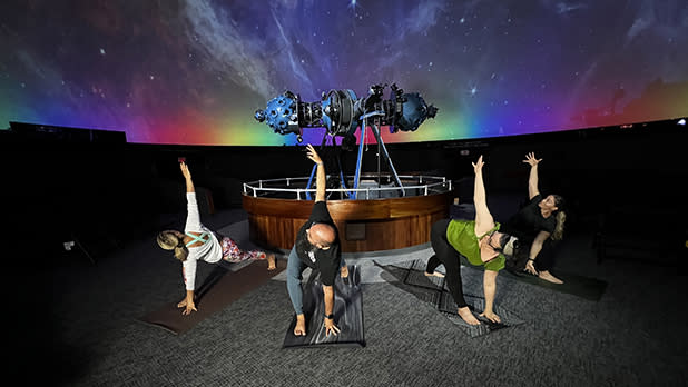 Four people practicing yoga in front of the Strasenburgh Planetarium's Mark Zeiss VI projector, with a rainbow and star-filled sky on the Planetarium dome.