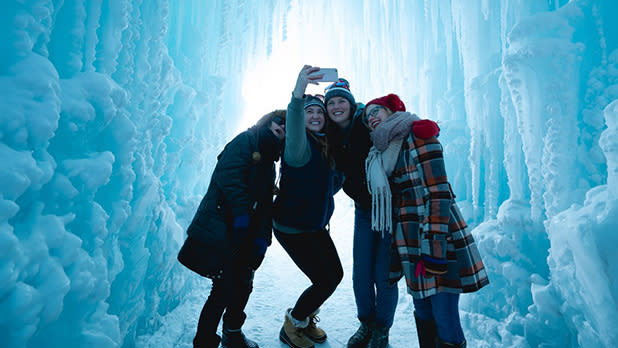 A group of young women in winter clothing huddle together to take a selfie inside the blueish white ice castle entrance