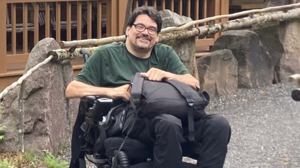 Jason Thurston, a man in a power chair wearing a green t-shirt, dark-framed glasses, in front of a wooden building at the International Paper John Dillon State Park