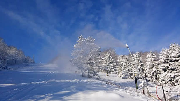 A snow machine is spreading snow across the slopes at Kissing Bridge with snow-covered trees in the background