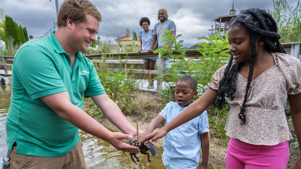 A Long Island Aquarium employee shows a crab to two children at the water's edge while two adults look on from the boardwalk