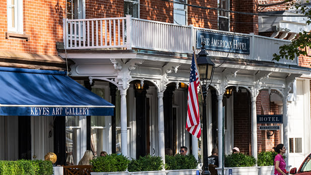 Street view of a historic brick building featuring "The American Hotel" sign in Sag Harbor. Blue canopy with "Keyes Art Gallery." US flag and outdoor seating.