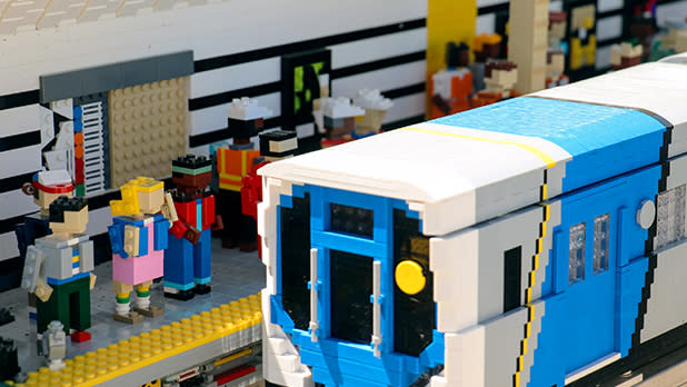Lego figurines stand on a platform outside a blue and white Lego brick subway train