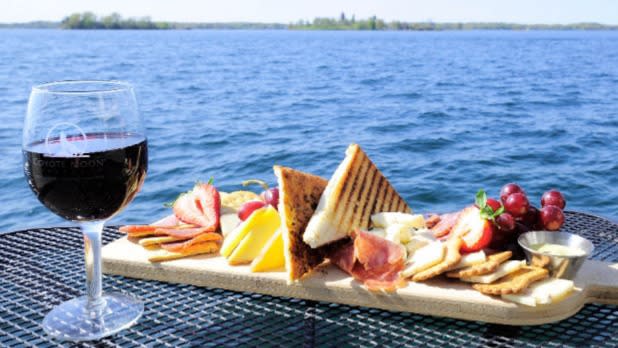 A glass of wine and a cheese plate on a table in front of a lake.