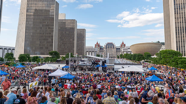 Crowd gathers in front of an outdoor stage for a concert at Empire State Plaza with the Capitol Building and The Egg performance center in the background.