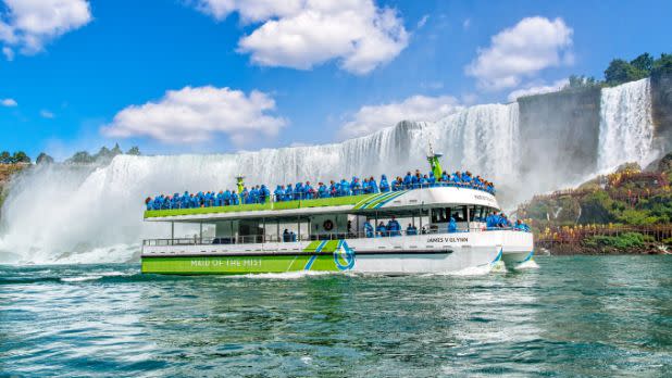 A group of people in blue raincoats aboard the green and white maid of the mist electric ship with views of the Niagara Falls behind it
