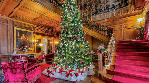 Lavishly decorated room with a large Christmas tree, red carpeted stairs, ornate wooden paneling, and elegant furniture at the Staatsburgh State Historic Site.