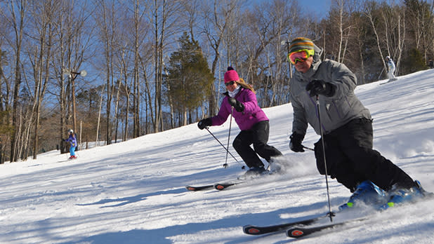 A woman in a purple jacket and black pants and a man in a gray jacket and black pants are skiing down the hillside at Mount Peter