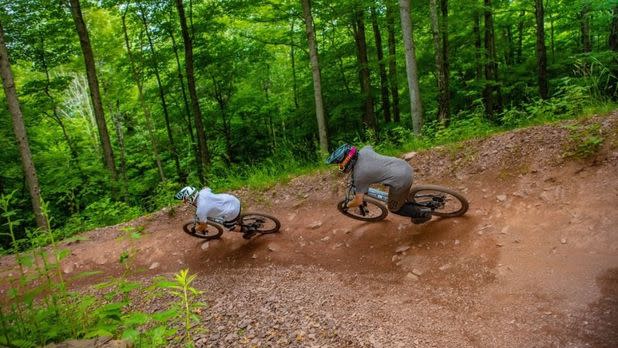 Two people mountain biking down a dirt road surrounded by the bright green forests of Windham Mountain