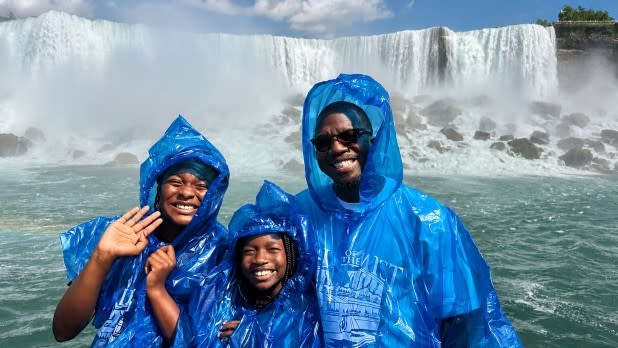 A family wearing blue rain ponchos aboard the Maid of the Mist boat tour smiles in front of a powerful waterfall at Niagara Falls. The sky is partly cloudy, enhancing the vibrant scene.