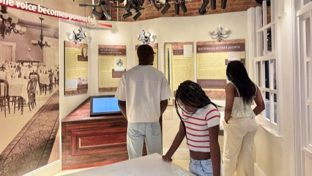 Three people view an exhibit with information panels and historical photos at the Niagara Falls Underground Railroad Heritage Center. The display discusses the role of African Americans in history. The scene is educational and engaging.