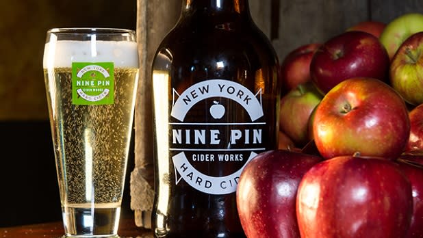 Glass of cider next to a brown growler and stack of red apples