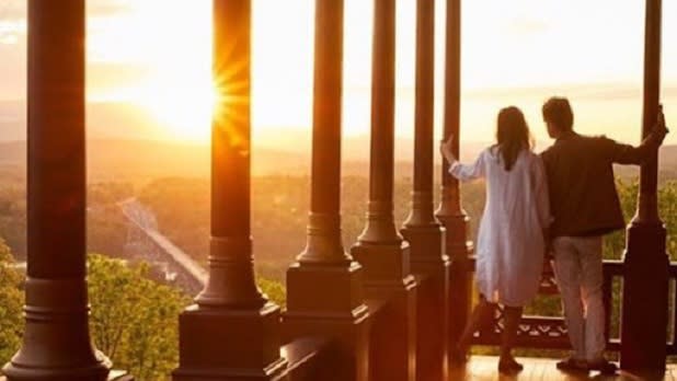 A woman in a white dress and a man in a brown jacket and khaki pants looking out at the sunset from an outdoor patio