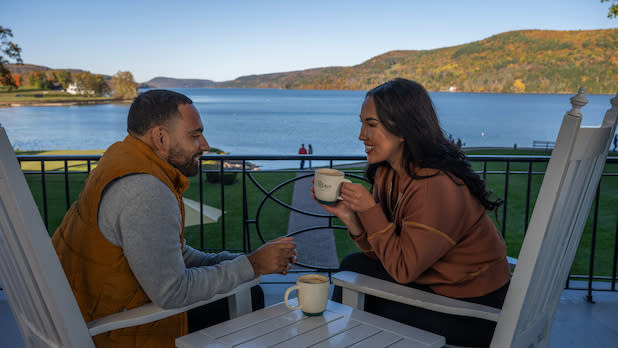 a man and a woman sip from mugs in rocking chairs looking out a Otsego Lake and fall foliage