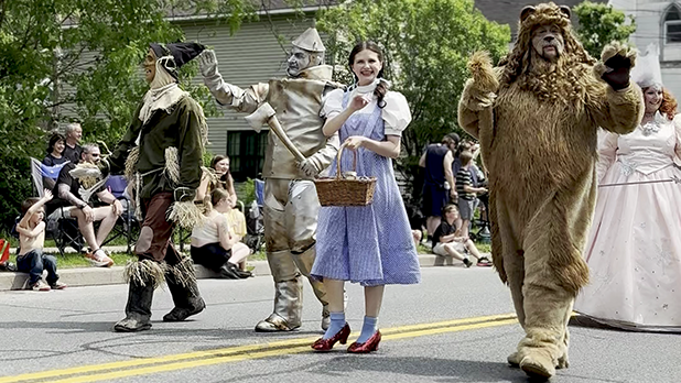A parade features characters resembling those from "The Wizard of Oz" walking joyfully at Oz-Stravaganza. Dorothy leads with a basket, followed by the Scarecrow, Tin Man, and Cowardly Lion.
