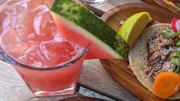 A pink cocktail with a slice of watermelon stands on a table next to a plate of tacos at Casa Enrique