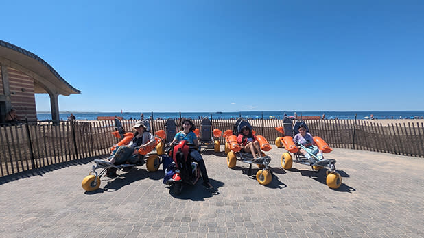 Four people in beach wheelchairs and one in a motorized scooter on stone pavement at Brighton Beach
