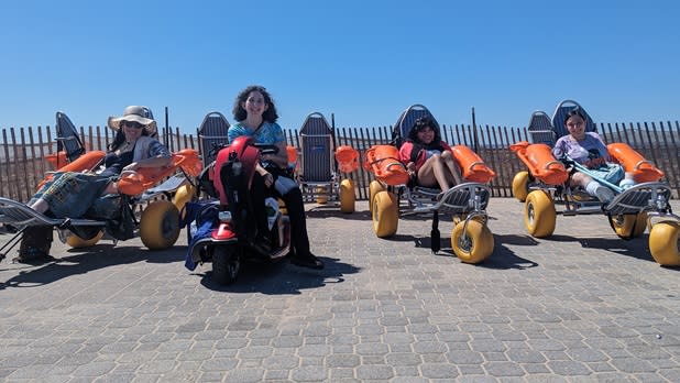 Three people in beach wheelchairs and one in a motorized scooter on stone pavement at Brighton Beach