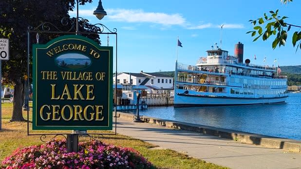 a green sign that reads "welcome to the Village of Lake George" in gold lettering with a view of the lake and a historic steamboat in the background