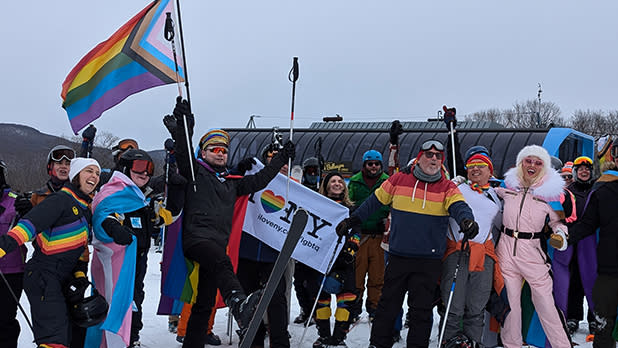 A joyful group of skiers in colorful rainbow and Pride-themed outfits hold flags, celebrating unity and diversity on a snowy slope with overcast skies during Catskills Pride Ski Weekend at Belleayre Mountain.