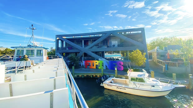 A modern building with large 'FIPINES' letters in front, seen from a dock with boats and a ferry under a clear blue sky.