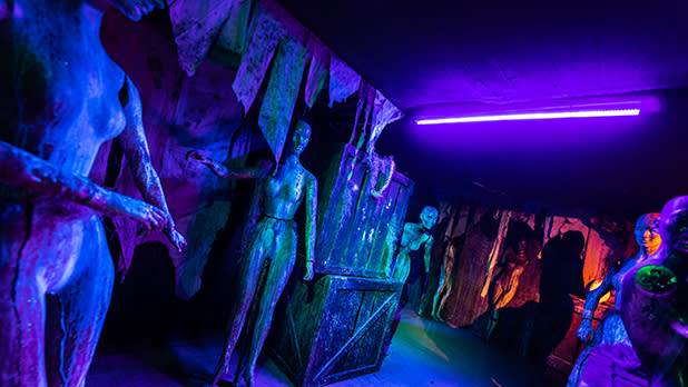 An eerie UV-lit cave attraction at Field of Horrors in Troy features life-size figures and jagged rock formations in vivid blue, purple, and green, creating a surreal, spooky mood.