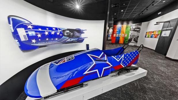 Blue Team USA bobsled with white outlined stars and red stripes on display at the Lake Placid Olympic Center in front of a large photo of a Team USA bobsled
